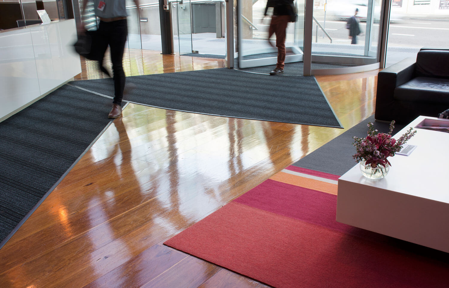 Custom shaped entrance mats are laid in front of revolving doors. People in work clothes walk over the top.